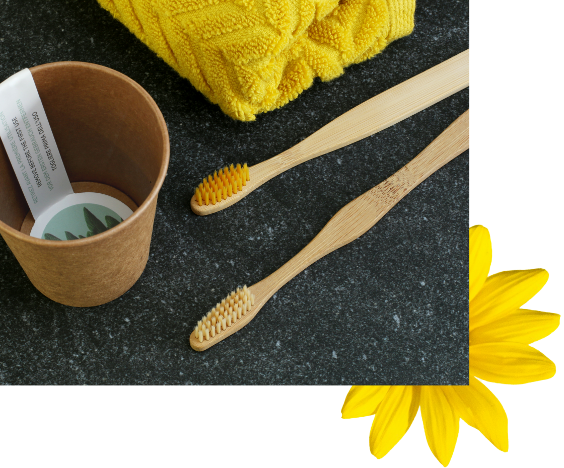 A pair of bamboo toothbrushes rest next to a brown paper container, with a bright yellow towel and a daisy in the corner. The scene has a clean, minimalist aesthetic.