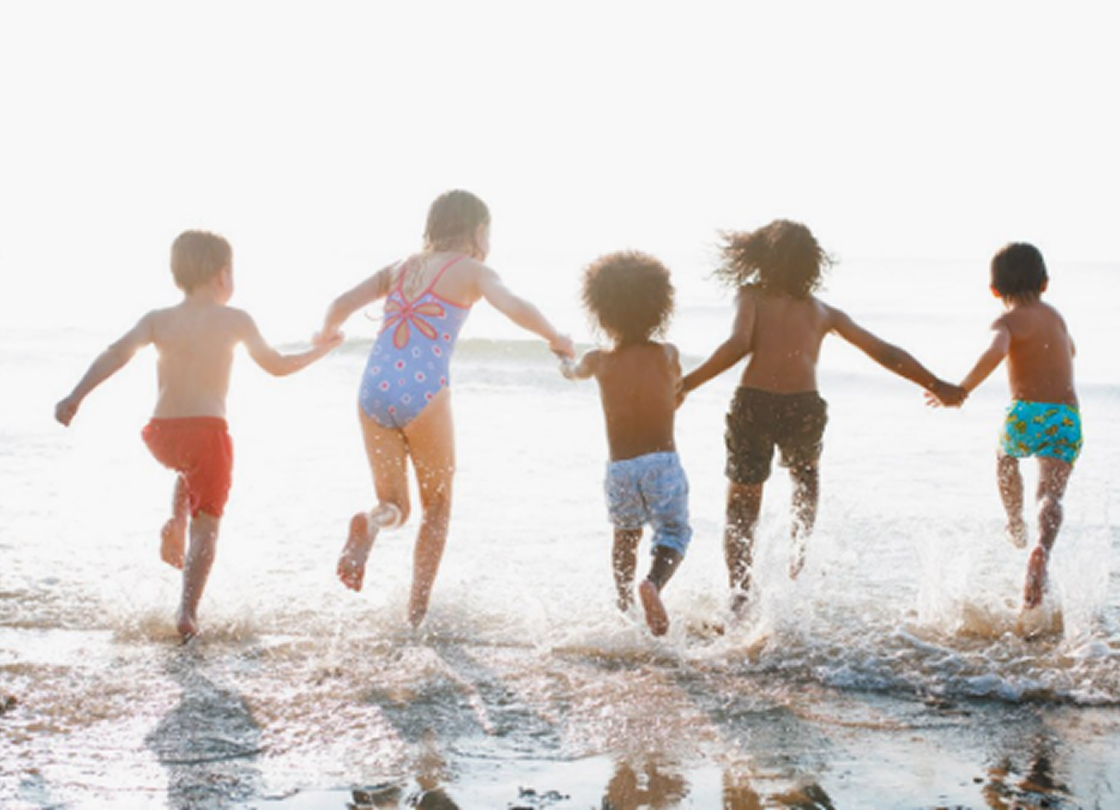 A group of five children, holding hands, joyfully wade into the ocean, splashing water as they run along the shore. They are backlit by the sun, creating a bright, cheerful atmosphere.