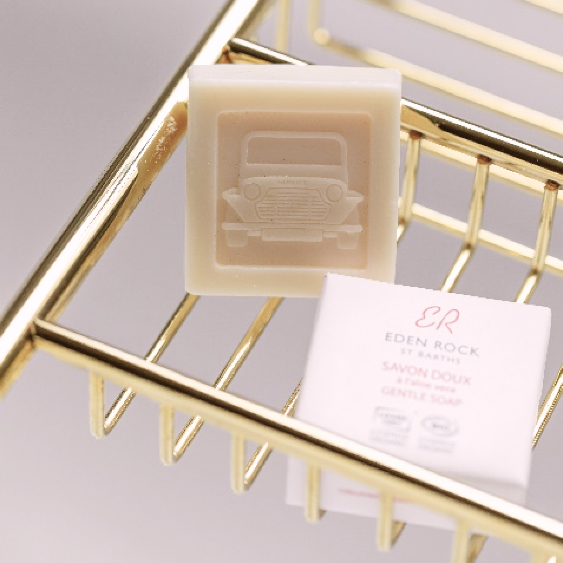 A bar of light-colored soap with a vintage car design sits on a gold wire soap dish beside a small, white box labeled "Eden Rock" gentle soap.