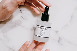 A hand holds a frosted glass dropper bottle labeled "CODAGE" against a marble surface.
