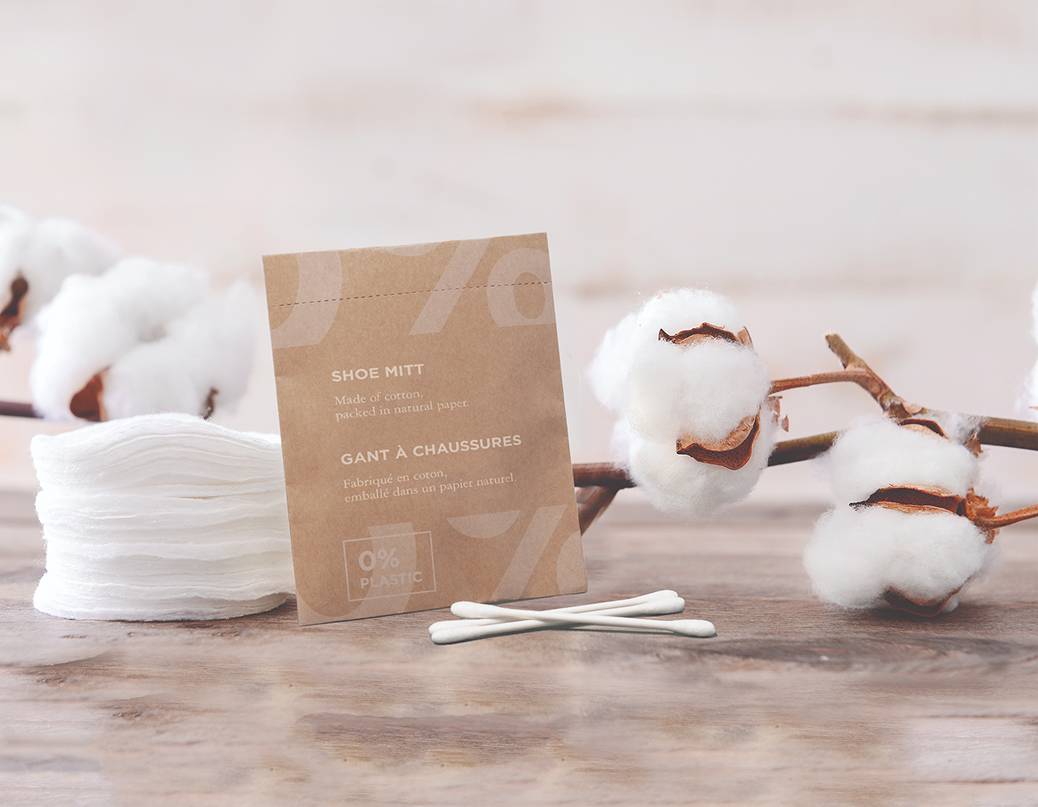 A package of shoe mitts made from natural cotton, surrounded by cotton bolls and a few cotton swabs. The background features a wooden surface.