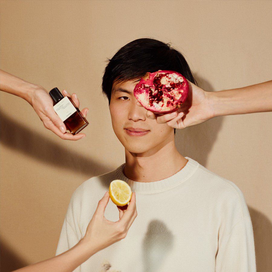 A young man with dark hair is centered against a beige background, flanked by two hands holding a bottle of perfume, a pomegranate half, and a lemon. The arrangement creates a vibrant and artistic display.