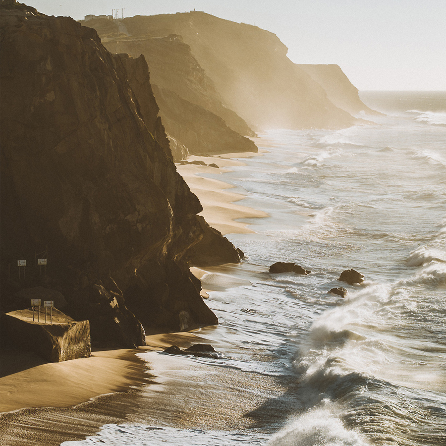A rugged coastline with steep cliffs meets the ocean, where waves crash against the shore in a misty, sunlit atmosphere. The scene captures the natural beauty and power of coastal landscapes.