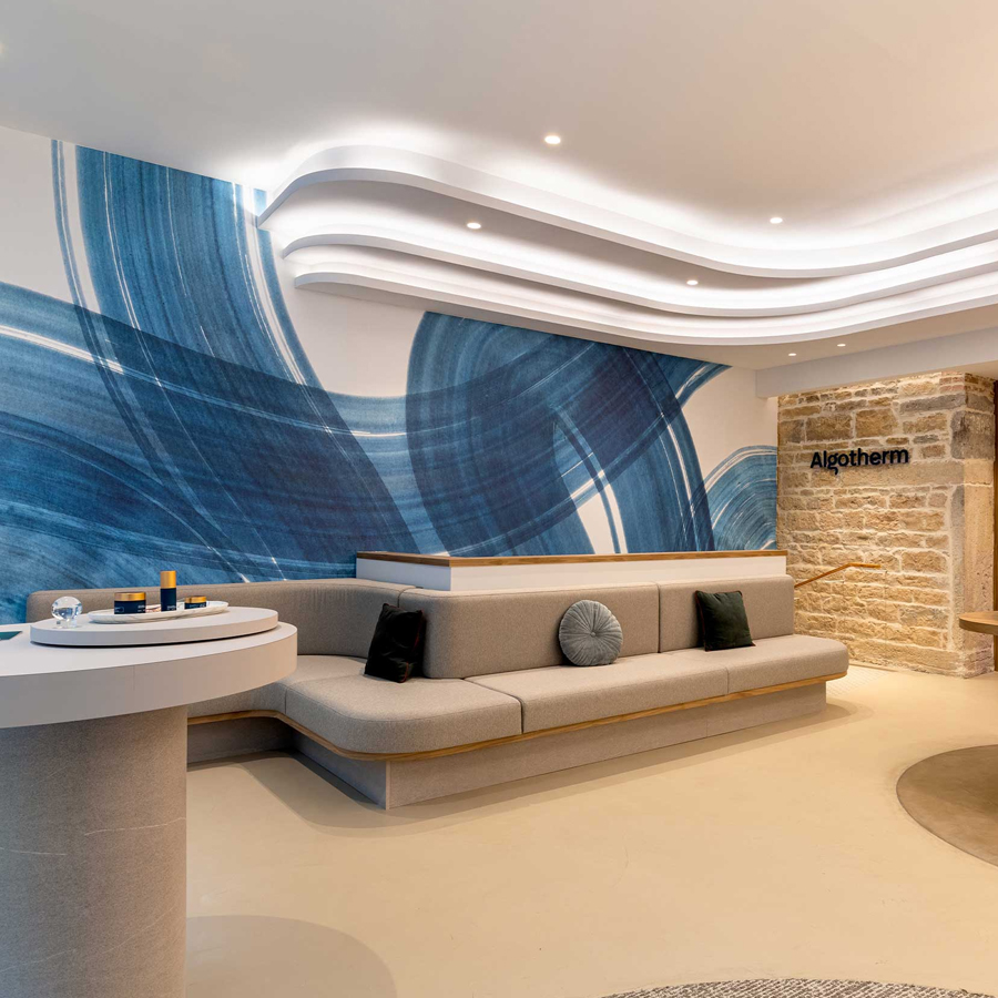 A modern reception area featuring a curved gray sofa, circular white table, and a blue abstract mural on the wall, with branding for "Algotherm" displayed prominently. Soft lighting enhances the contemporary design.