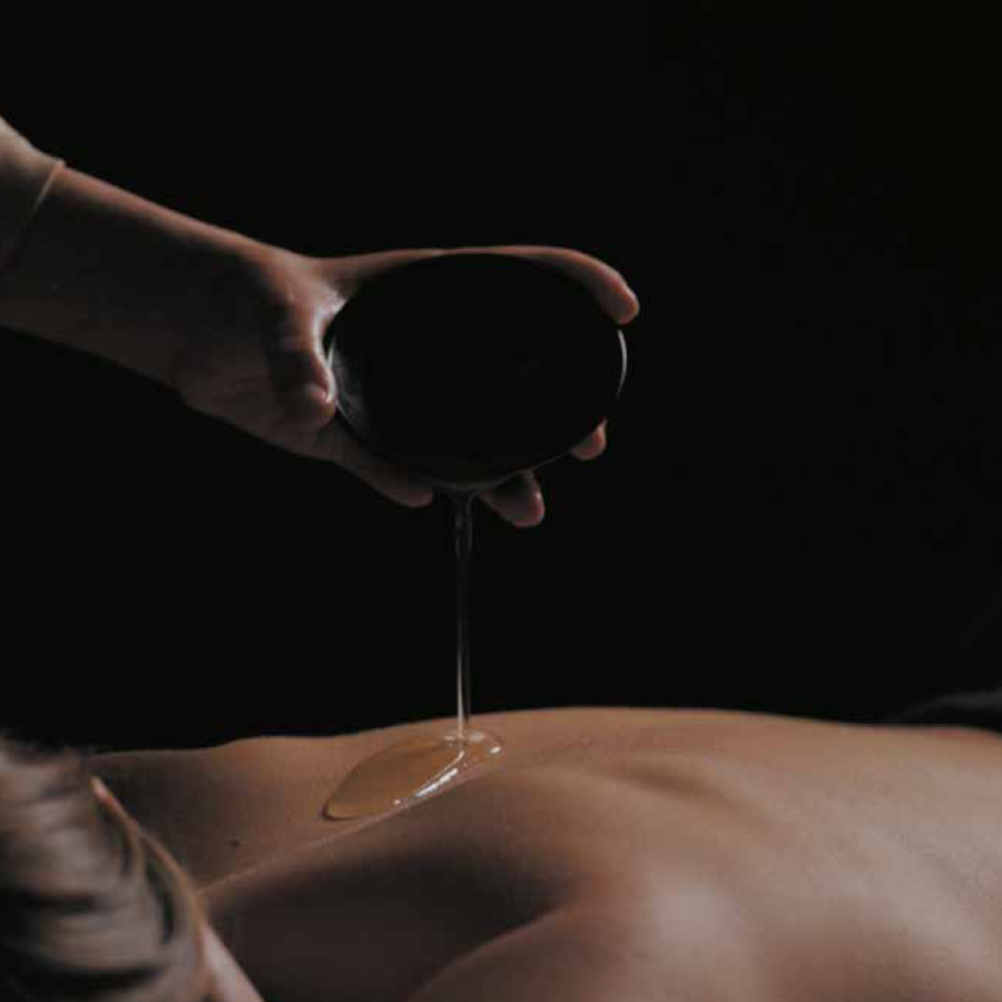 A hand pouring massage oil onto the back of a person lying on a massage table, set against a dark background.