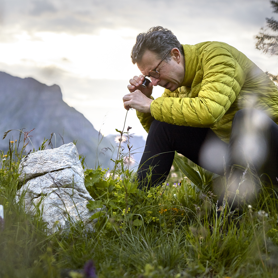 A person in a yellow jacket kneels beside a rock, examining a plant closely with a magnifying glass in a scenic outdoor setting.