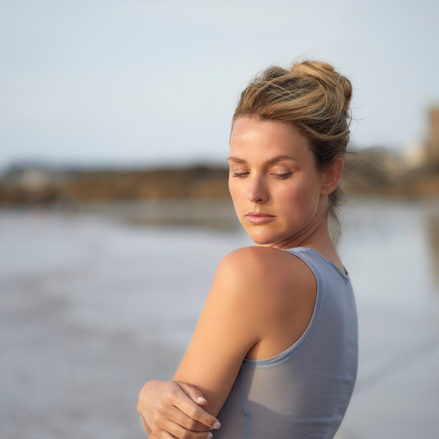 A woman with a bun hairstyle stands by the beach, looking serene with her eyes closed, wearing a gray tank top. The ocean and soft sand are blurred in the background.