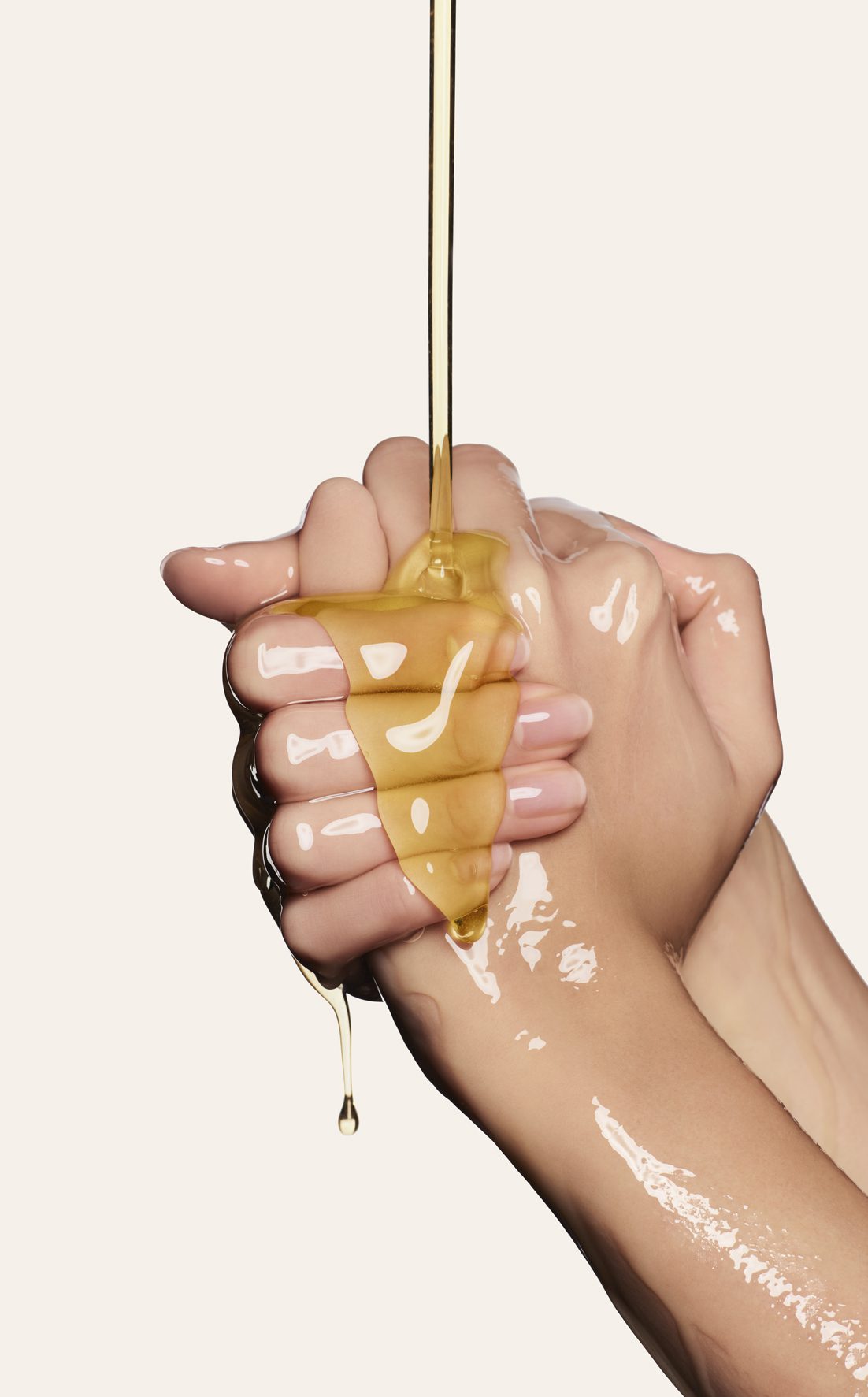 A close-up of two hands holding a dripping piece of honey or golden viscous liquid, emphasizing the texture and sheen. The background is a soft neutral color.