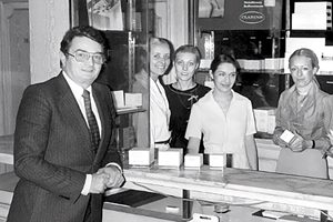 A group of five people, including a man in a suit and four women in professional attire, stand behind a counter showcasing various products in a store setting. The environment appears to be a cosmetic or beauty-related shop.
