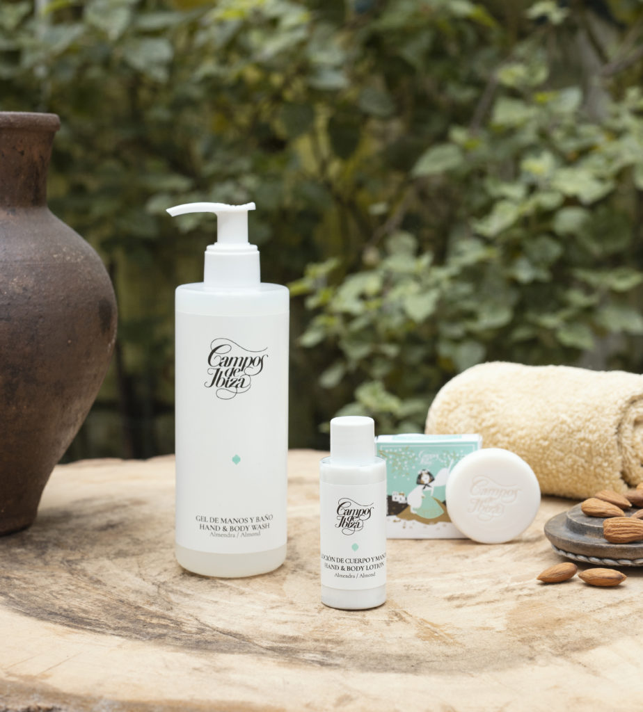 A collection of beauty products, including a large pump bottle, a small bottle, and a soap bar, displayed on a wooden table with a clay pot and almonds in the background. Green foliage creates a natural setting.