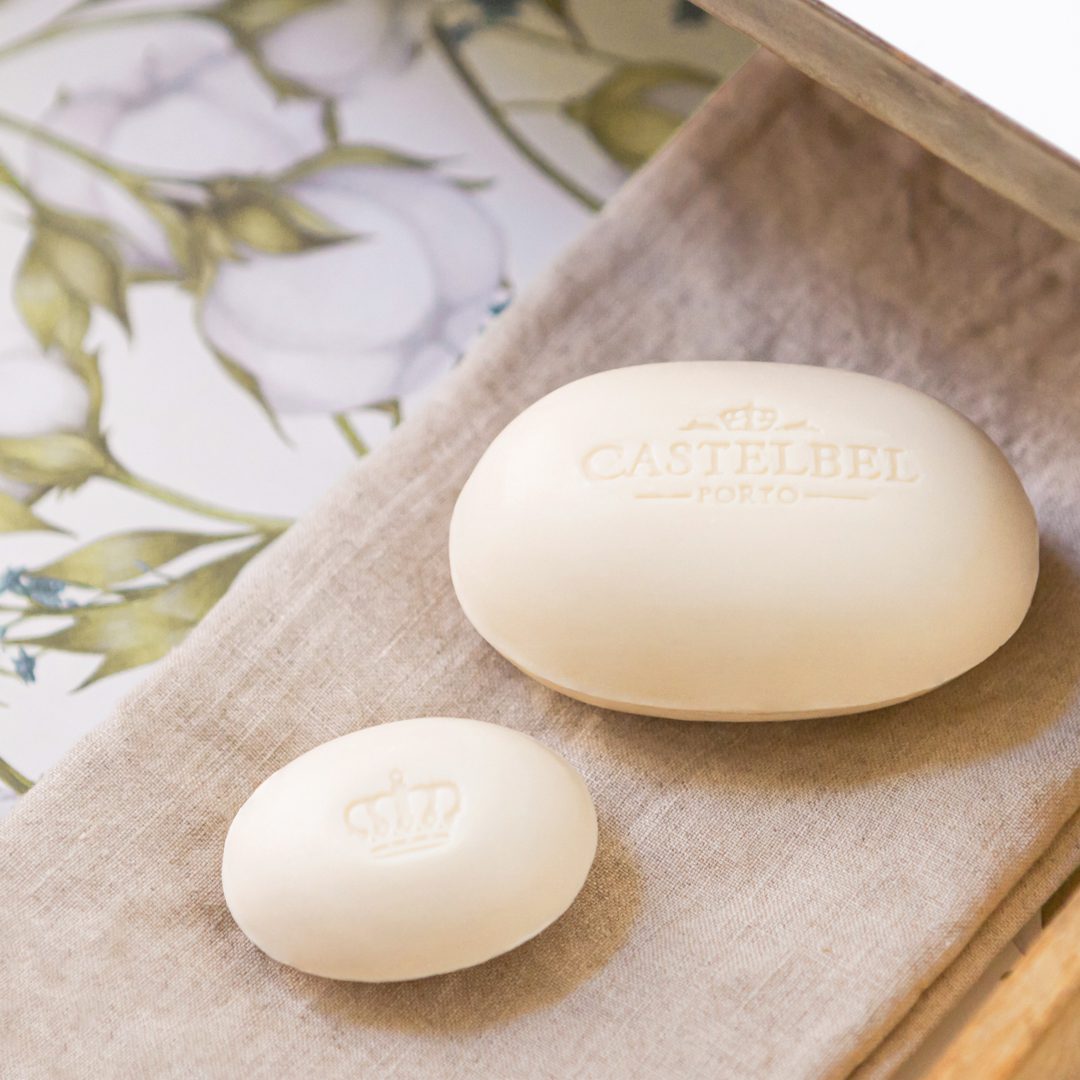 Two cream-colored soaps are arranged on a neutral fabric, with one larger soap displaying the embossed text "CASTELBEL PORTO" and a smaller soap featuring a crown design. The background includes a floral pattern.