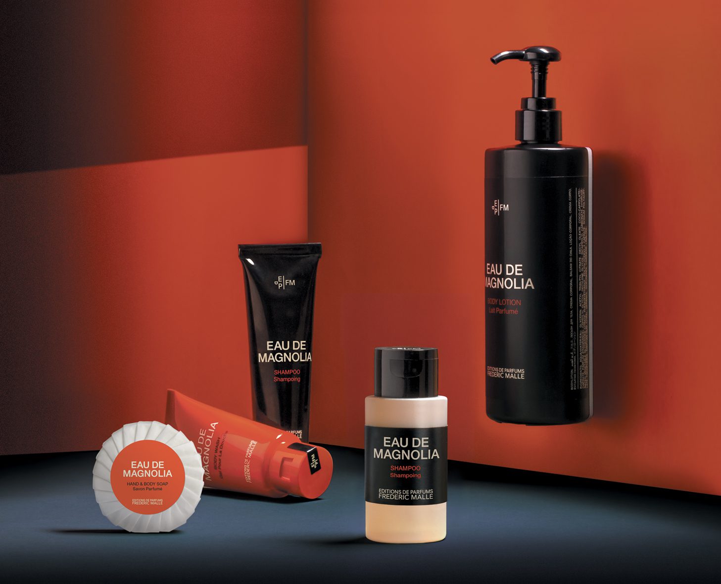 A collection of Eau de Magnolia personal care products displayed against a contrasting red and black background, including a body lotion, shampoo, and cream.