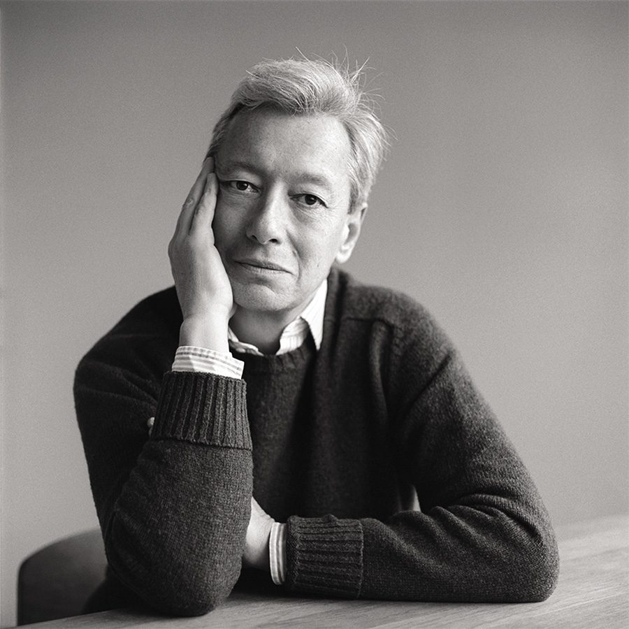 A contemplative man with gray hair rests his chin on his hand while sitting at a table, wearing a dark sweater and a collared shirt. The photo is in black and white, highlighting his thoughtful expression.