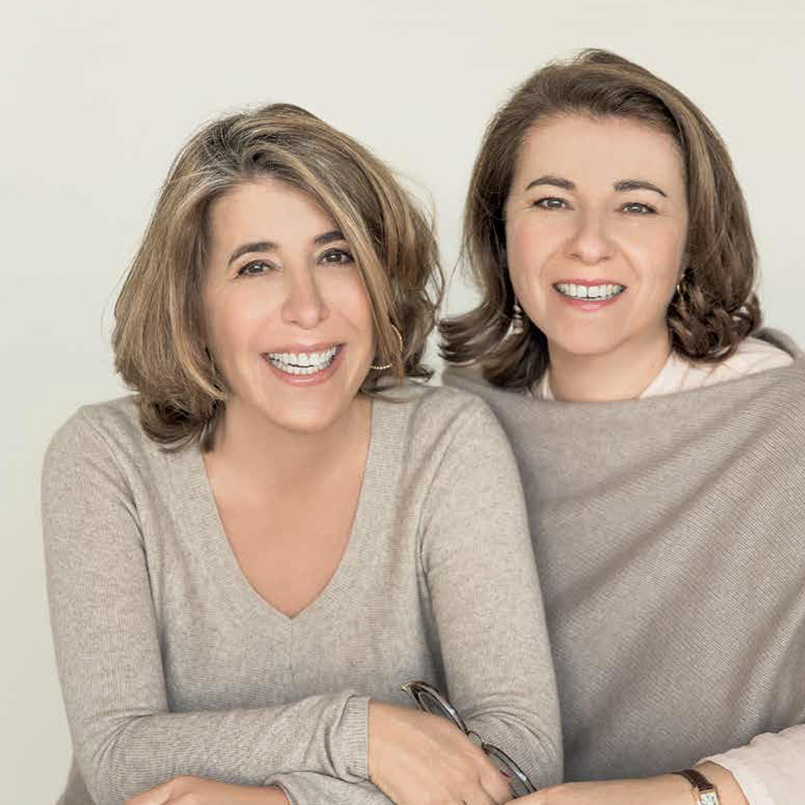Two smiling women sit closely together, both dressed in light, neutral-colored sweaters against a soft background.