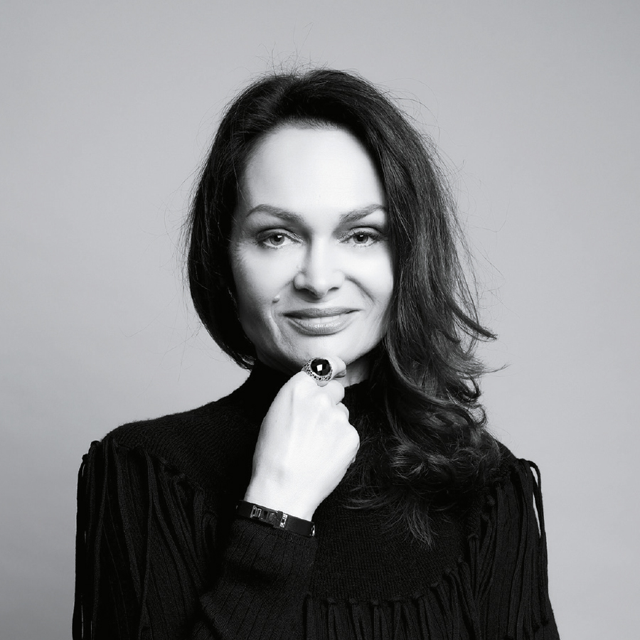 A woman with long, wavy hair wearing a black turtleneck and a statement ring, smiles confidently against a light grey background.