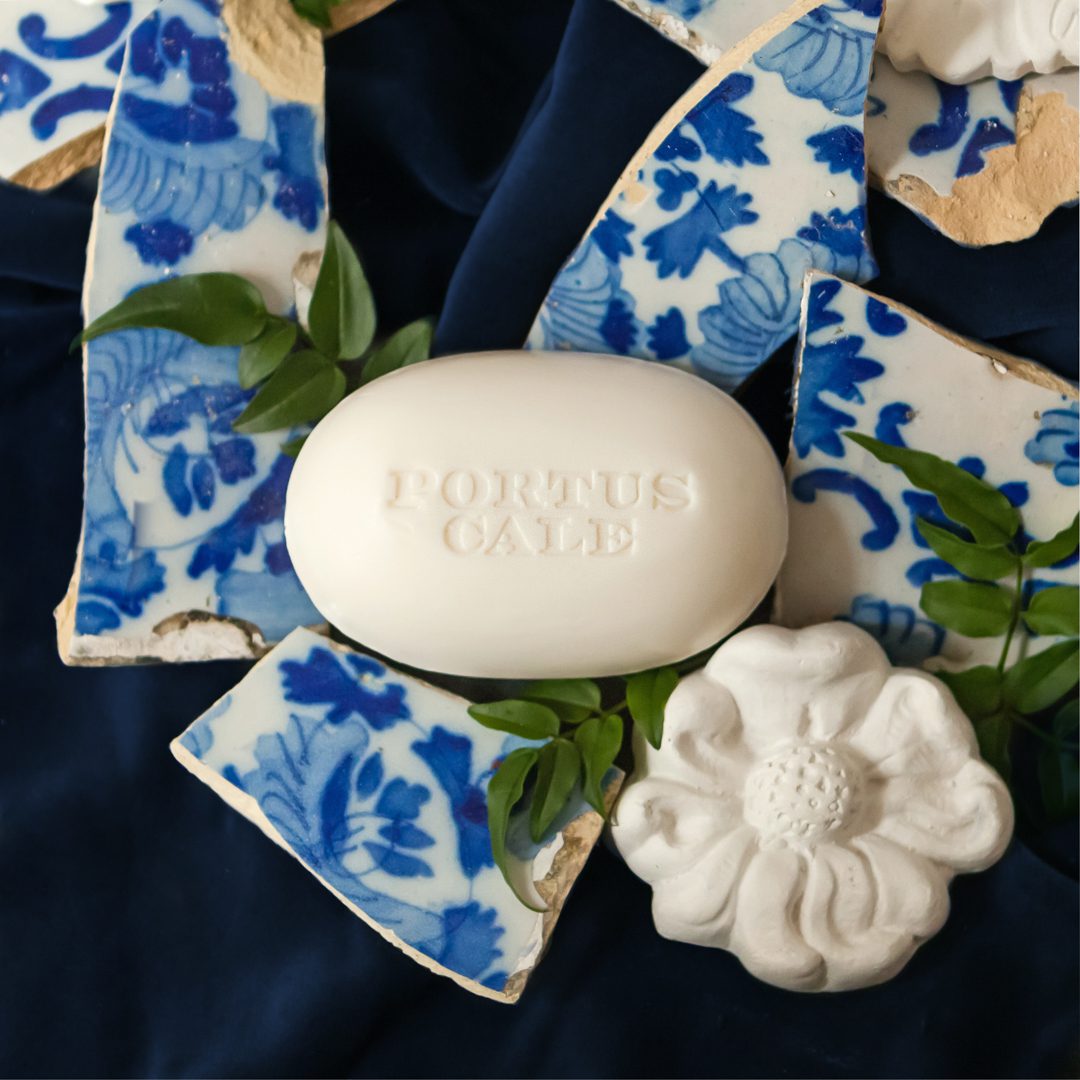 A white soap bar embossed with "PORTUS CALE" surrounded by blue and white pottery shards and a white flower sculpture on a dark fabric backdrop.