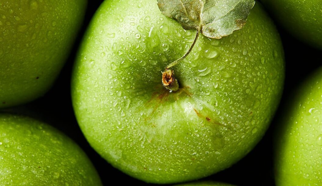 A close-up of a shiny green apple with droplets of water on its surface, surrounded by other apples. A small leaf is attached to the stem.