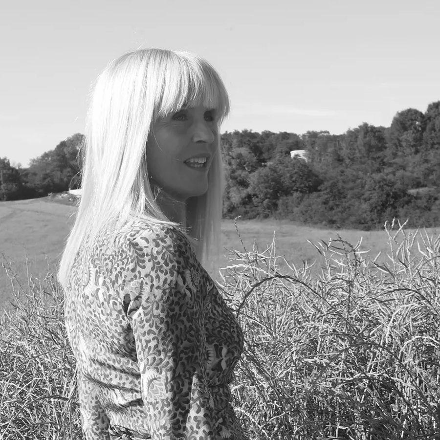 A woman with long blonde hair stands in a field, facing to the side, with greenery and trees in the background. She is wearing a patterned dress and appears to be smiling.
