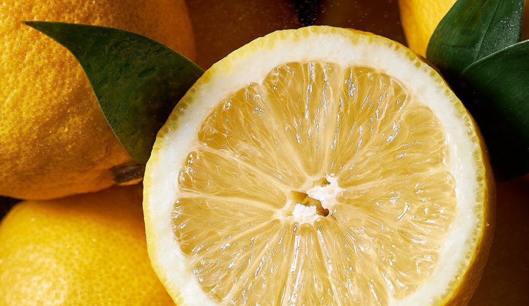 A close-up of a halved lemon, showcasing its juicy, textured interior, surrounded by whole lemons and green leaves.