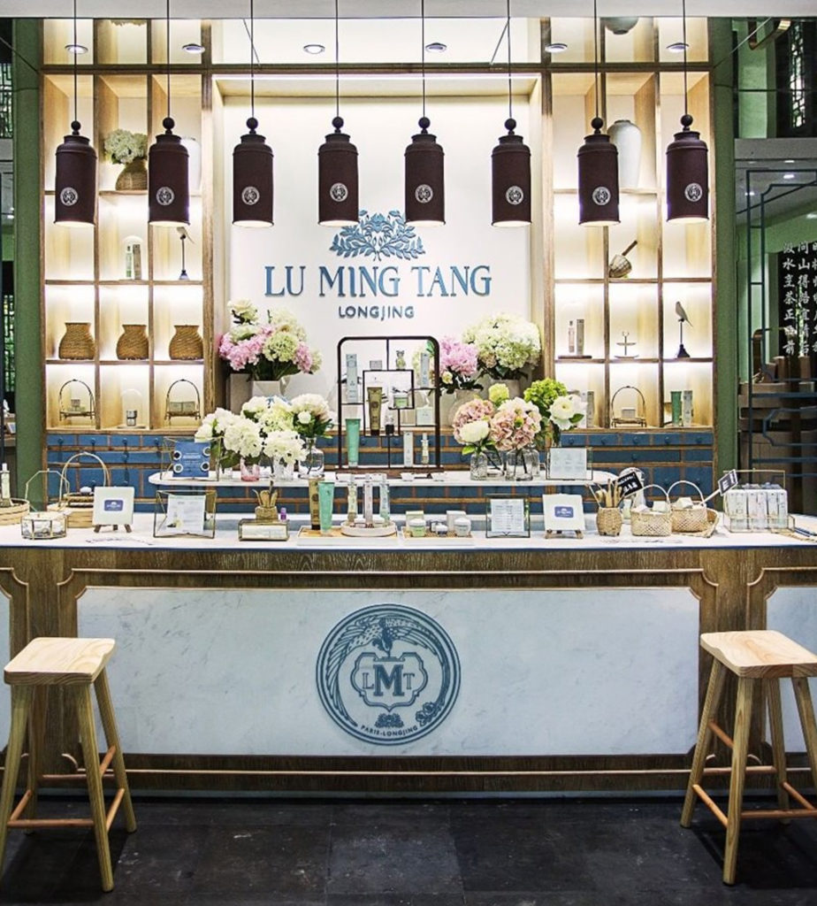 A beautifully arranged retail space for Lu Ming Tang, featuring elegant shelves adorned with decorative items and floral arrangements, alongside a display counter with various products. The overall aesthetic is modern and welcoming.