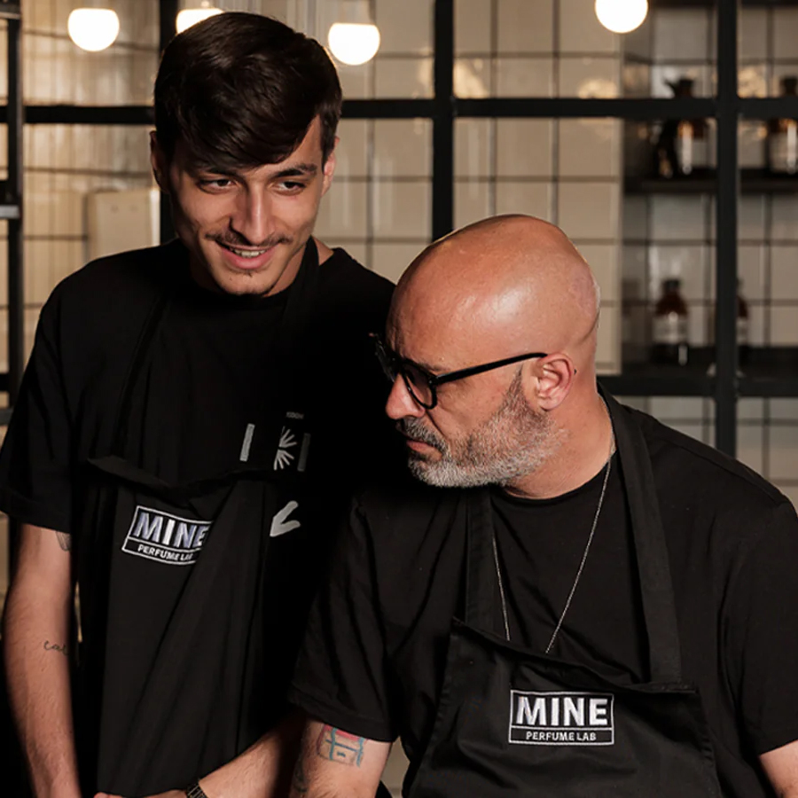 Two men in black aprons stand in a lab setting, smiling and engaged in conversation. The background features shelves with glass containers and warm lighting.