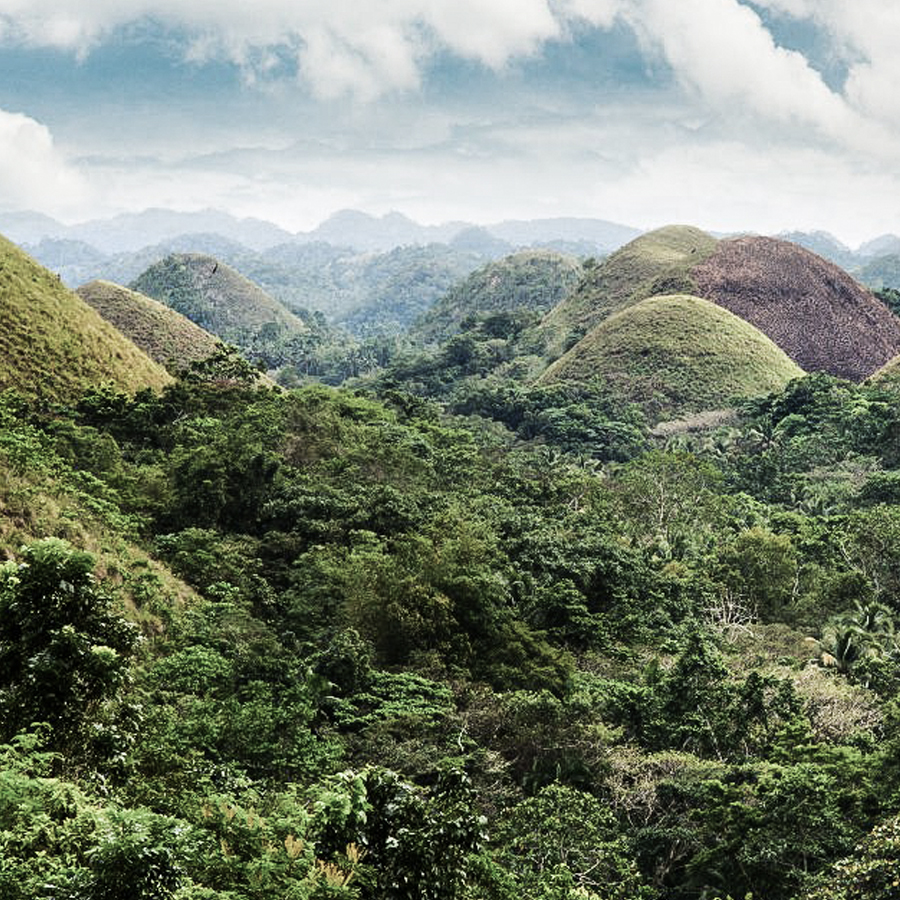 A lush green landscape featuring rolling hills and mounds, under a cloudy sky. The scene showcases dense vegetation and distant mountains.