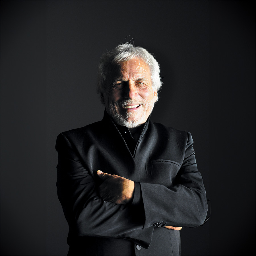 A smiling older man with white hair and a beard stands with his arms crossed, wearing a black jacket against a dark background.