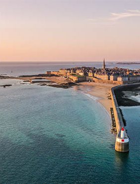 A coastal view of a town at sunset, featuring a sandy beach, rocky shoreline, and a lighthouse at the end of a jetty. The tranquil sea reflects warm colors of the sky.