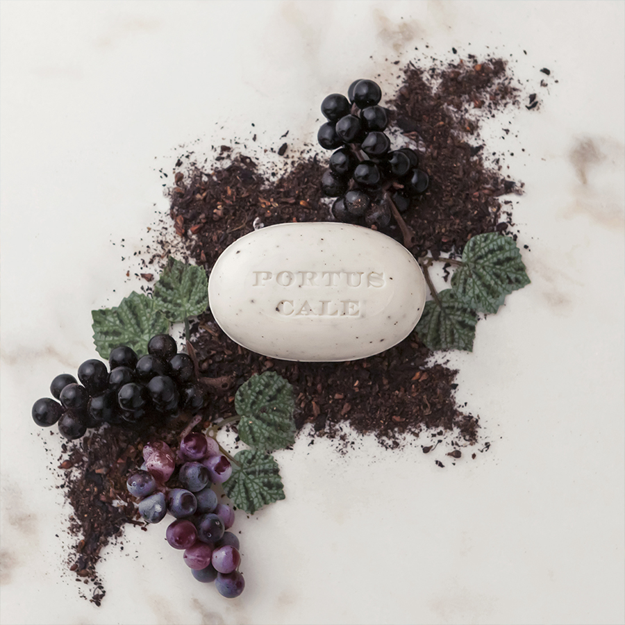 A white soap bar engraved with "PORTUS CALE" is placed among dark tea leaves and clusters of black and purple grapes, surrounded by green leaves.