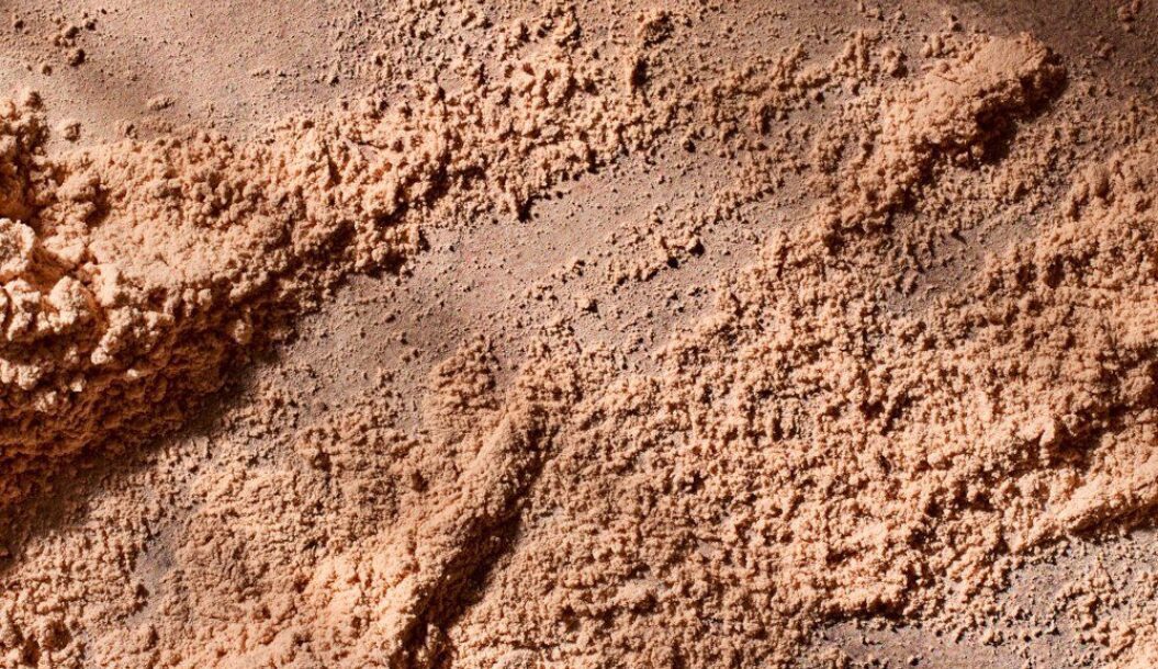 A close-up view of finely textured, reddish-brown powder resembling sand or soil, with uneven surfaces and subtle shadowing.