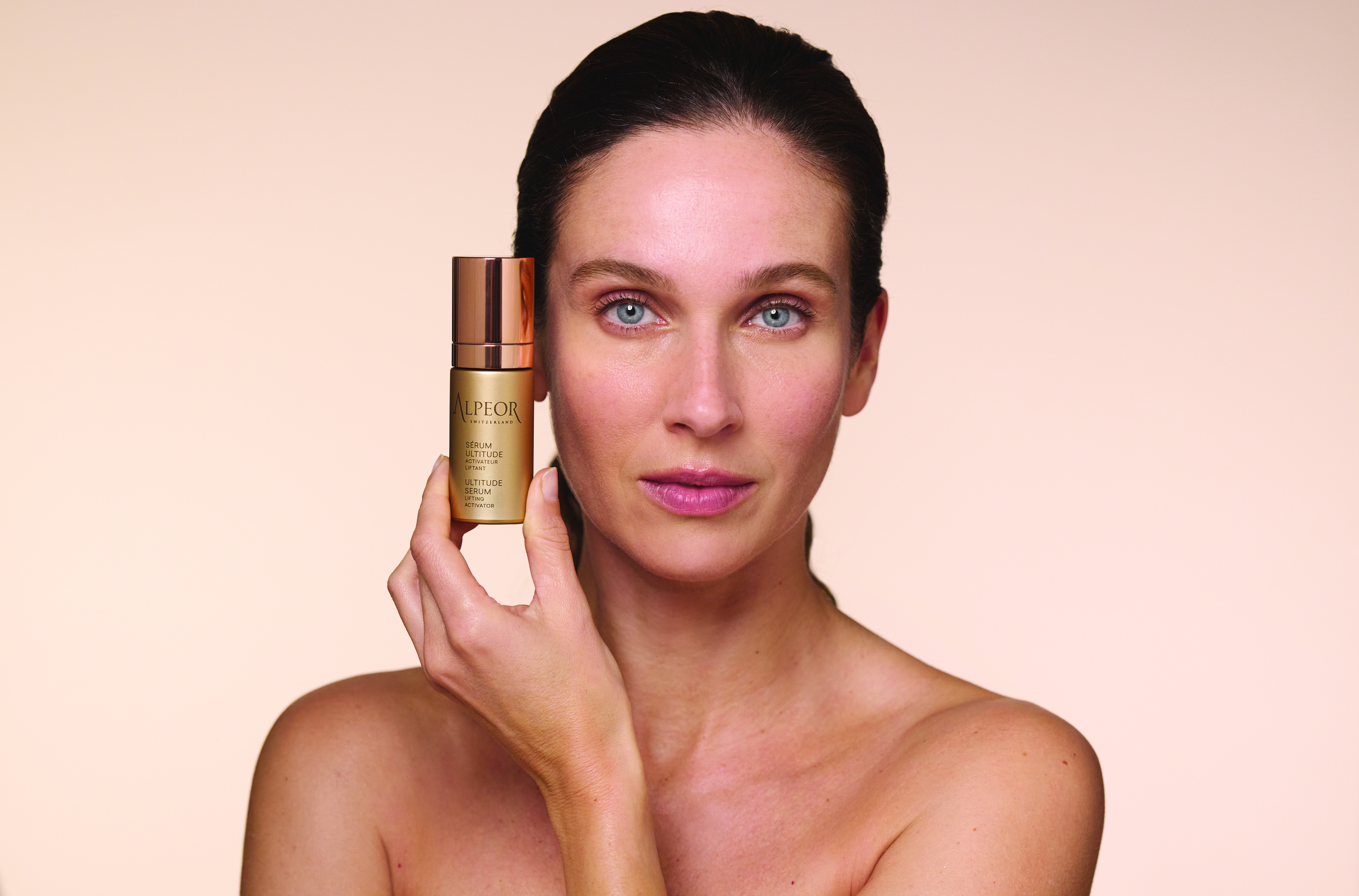 A woman with long dark hair holds a bottle of skincare product near her face, showcasing her radiant skin against a soft, neutral background.