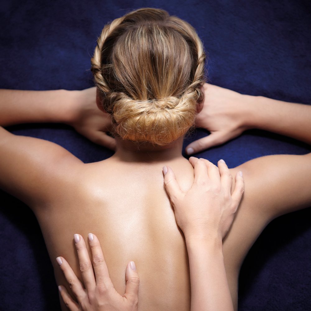 A person with long, braided hair is lying face down, receiving a back massage. Another hand is gently applying pressure to the shoulder area.