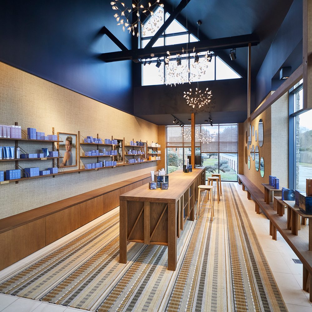 A modern retail space with a wooden counter, shelves displaying blue products, and large windows letting in natural light. The decor features contemporary lighting and a textured wall.
