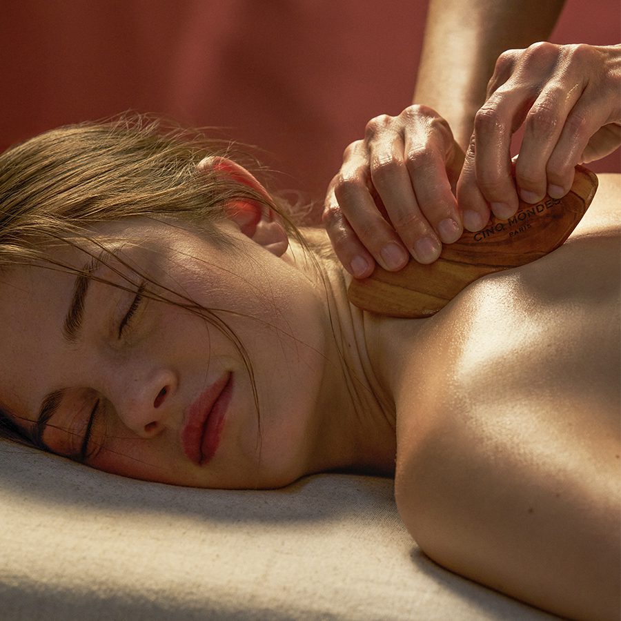 A person receives a back massage using a wooden massage tools, while lying on a soft surface with their eyes closed, displaying a relaxed expression. The setting has warm, soft lighting.