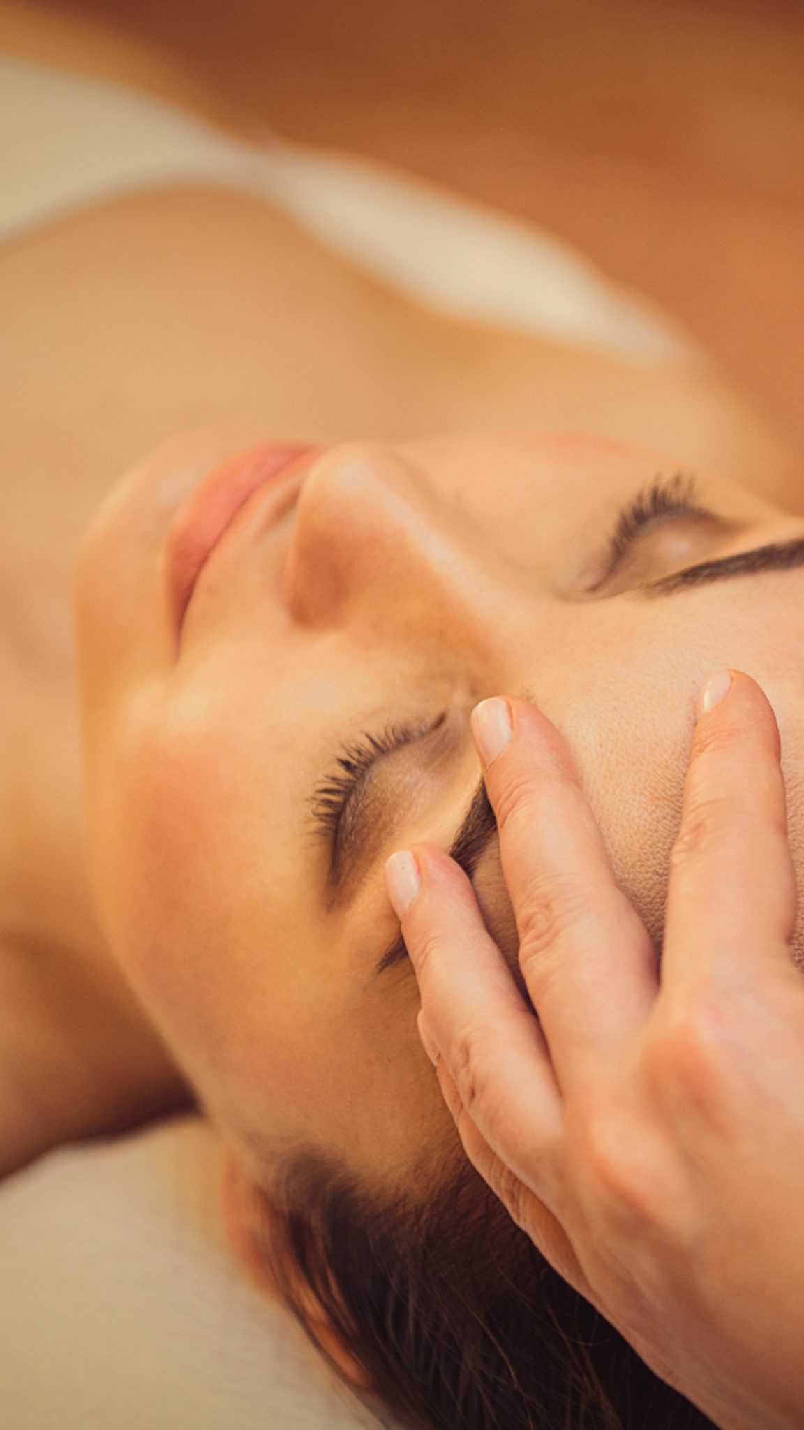 A woman is lying on her back with her eyes closed, receiving a soothing head massage. A hand gently rests on her forehead, creating a relaxing atmosphere.