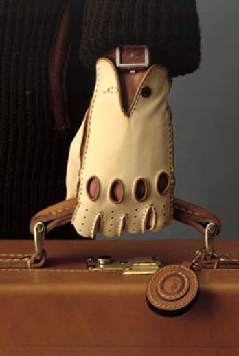 A close-up of a hand wearing a cream-colored driving glove holding the handle of a brown leather briefcase. The glove features fingerless design elements and decorative stitching.