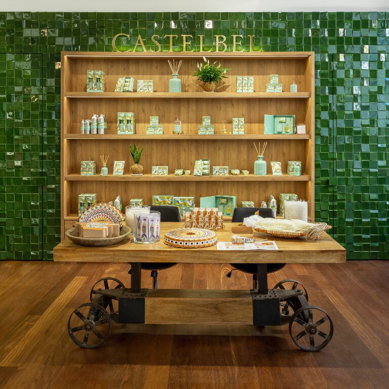 A wooden display table in front of a green tiled wall showcases various Castelbel products, including soaps, candles, and decorative items, arranged on a wooden shelf behind.