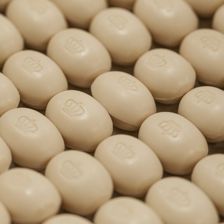 A close-up of neatly arranged beige oval-shaped candies, each embossed with a crown logo. The candies are displayed in a repetitive pattern on a flat surface.