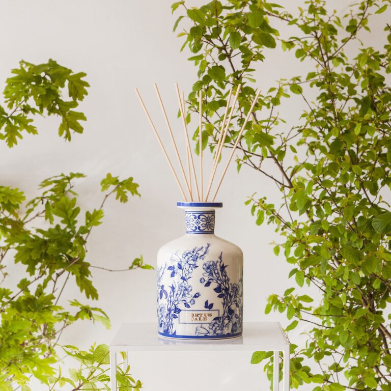 A decorative blue and white ceramic diffuser sits on a clear stand, with several bamboo reeds emerging from its neck, against a backdrop of lush green foliage.