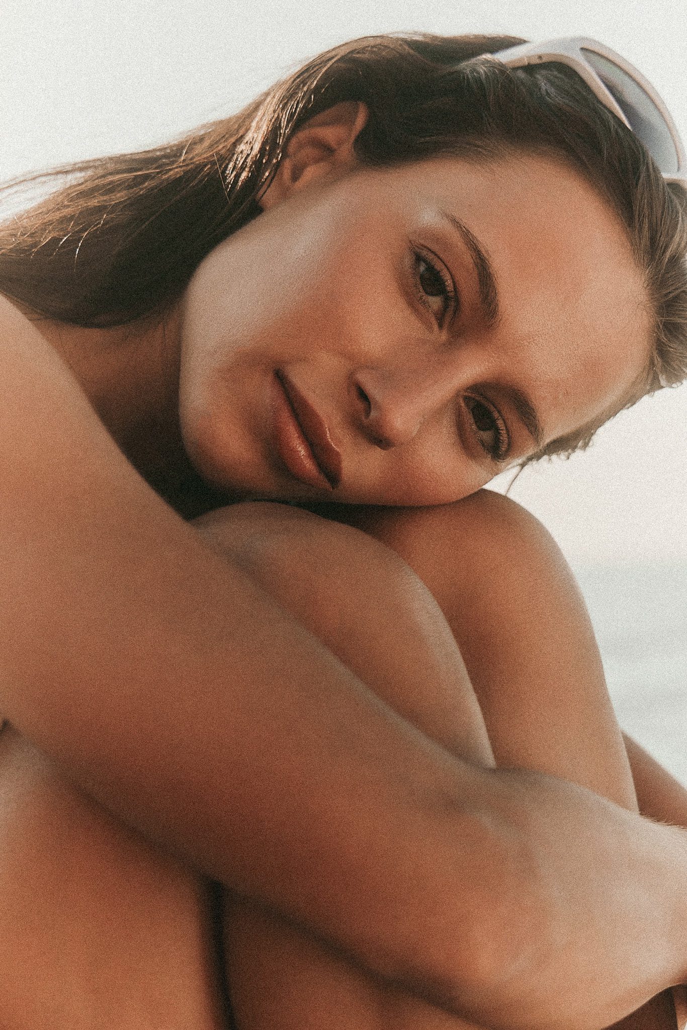 A close-up portrait of a woman resting her chin on her knees, gazing softly at the camera with a serene expression, wearing sunglasses in her hair and a neutral background.