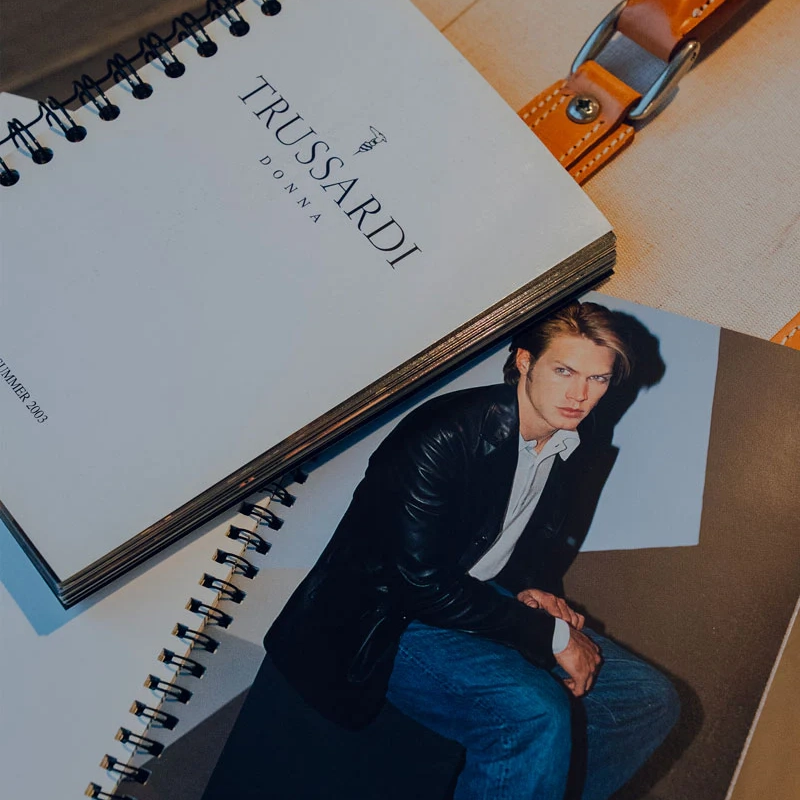 A stylish magazine featuring a model in a leather jacket is placed next to a spiral notebook labeled "Trussardi Donna." Both items are arranged on a light background with a leather strap visible.