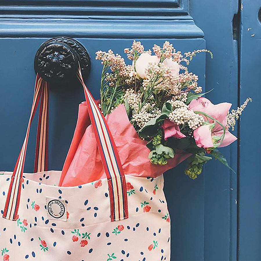A floral tote bag hangs on a blue door, with pink and white flowers peeking out, wrapped in colorful paper.