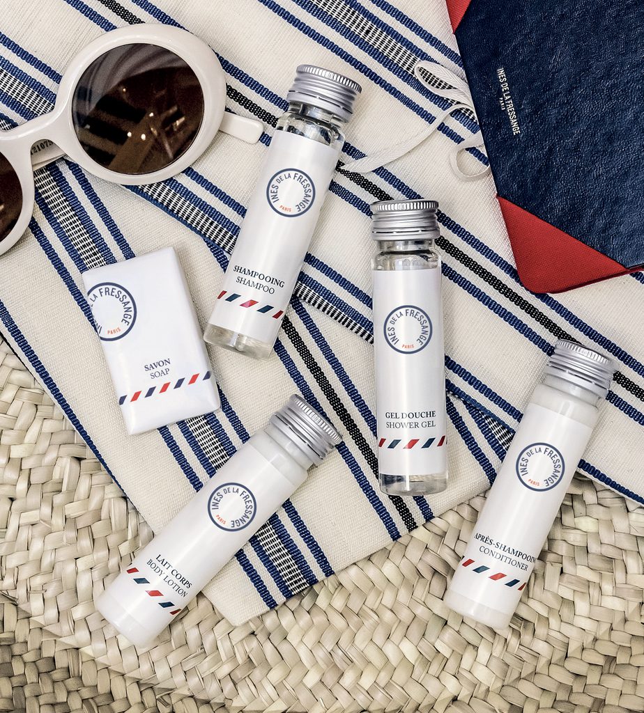 A collection of travel-sized toiletries, including shampoo, shower gel, hand soap, and conditioner, arranged on a patterned fabric with sunglasses and a notebook.