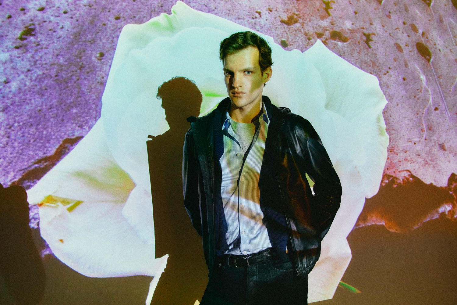 A person in a leather jacket stands confidently against a colorful projection of a large white flower. The background features soft, abstract colors and textures.
