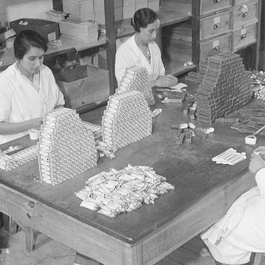Two women are seated at a wooden table assembling stacked packages, with various neatly arranged items and supplies around them in a workshop setting.