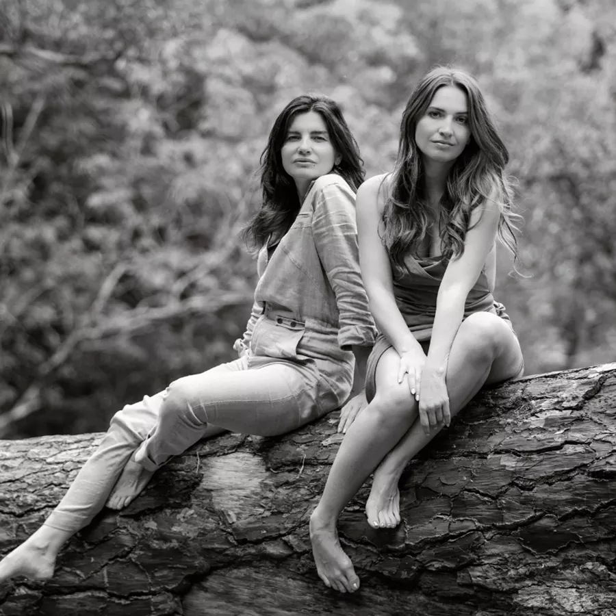 Two women sitting on a fallen log in a forest, both smiling and wearing casual clothing. The image is in black and white.