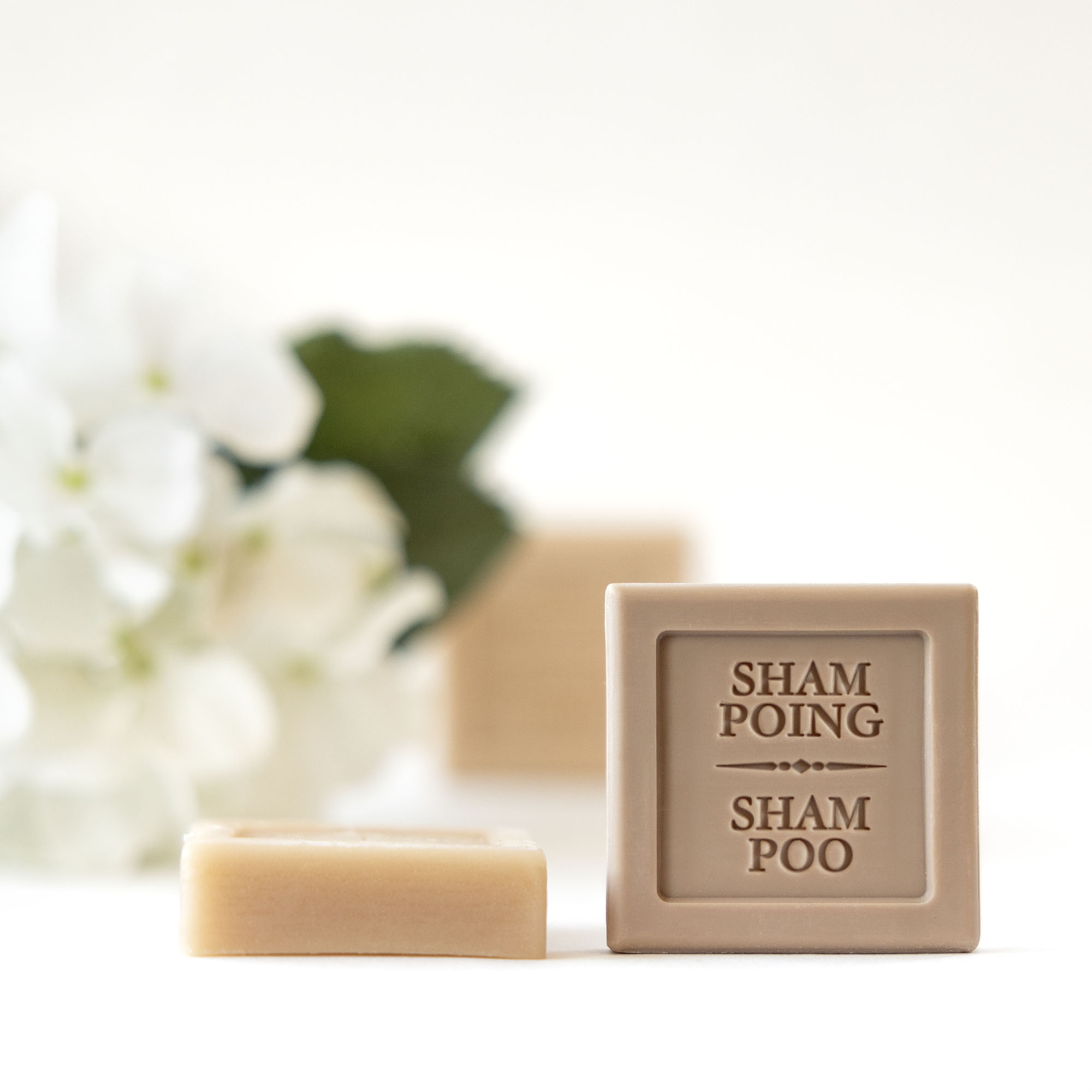 A bar of shampoo soap labeled "SHAMPOOING" beside a smaller rectangular piece, with soft white flowers and green leaves in the background.