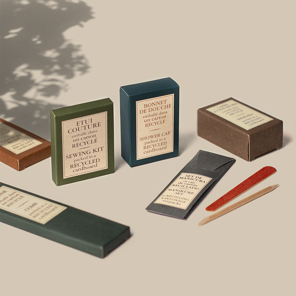 A collection of eco-friendly packaging boxes for various products, including a sewing kit and a shower cap, arranged artistically with wooden sticks and a shadow in the background. The boxes are made from recycled cardboard and feature minimalist labels.