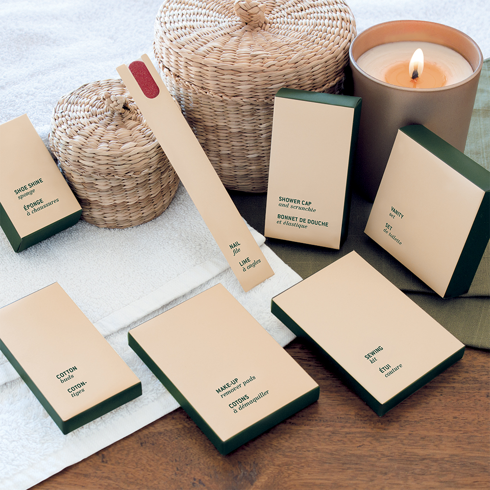 A laid-out collection of eco-friendly personal care items, including soap, shower cap, cotton pads, and a candle, arranged on a soft towel with wicker storage containers.