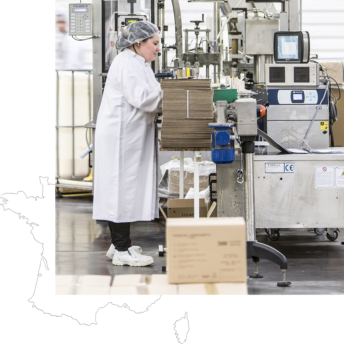 A worker in a white lab coat operates machinery in a packaging facility, surrounded by boxes and industrial equipment.